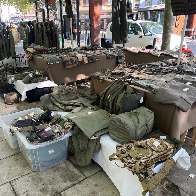 Vêtements militaires occasion, treillis et veste kaki exposés sur des tables dans un marché en plein air.